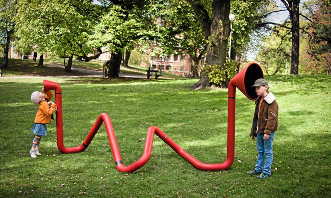 two children at either ends of a snaking tube in a playground. They are listening for eachother's voices.